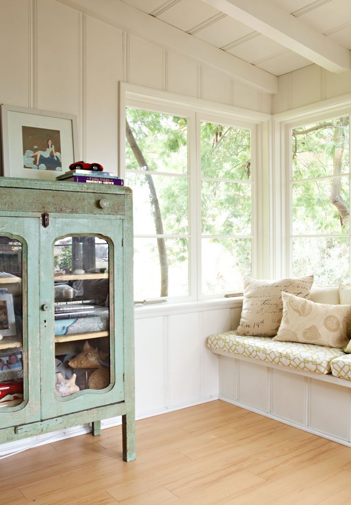 A vintage, soft green display cabinet in a loft with a light and bright window seat overlooking trees and nature outside. 