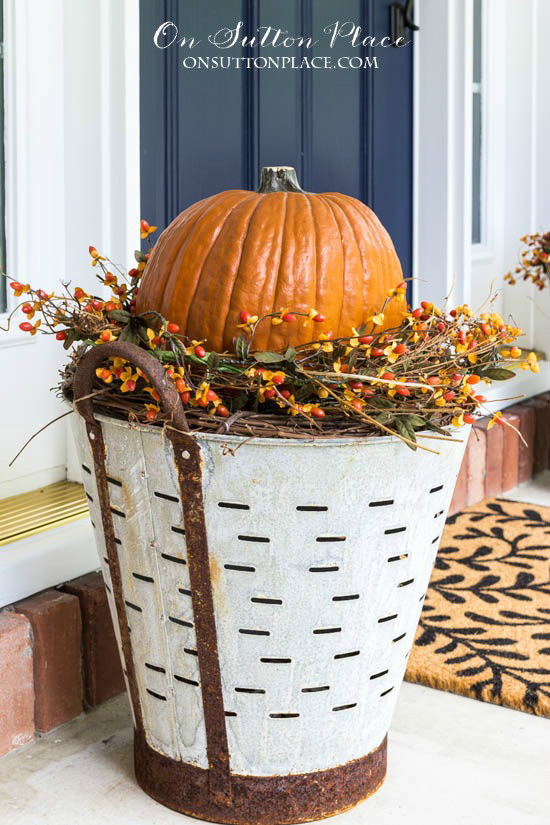 Rustic olive buckets filled with grapevines and a faux pumpkin, situated on a porch next to the front door. 