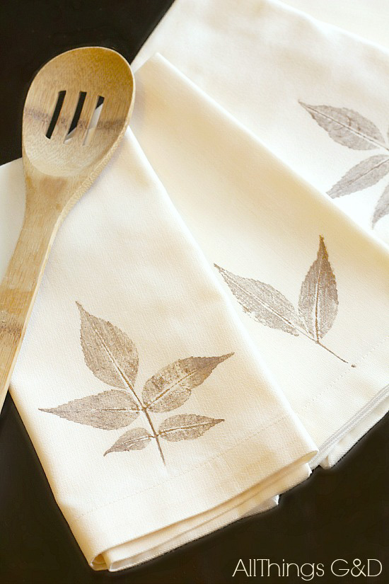 A set of four cream colored linen napkins each decorated with a hand stamped gold leaf and situated next to a slotted wooden spoon. 