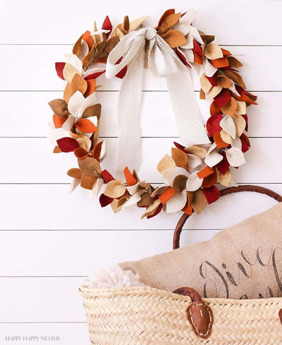 DIY fall craft, a hanging wreath covered in felt multicolored leaves in front of a white shiplap backdrop with a woven travel bag in the foreground. 