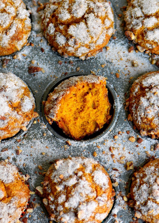 Metal muffin tin filled with pumpkin muffins and sprinkled with crunchy sugar, the middle muffin is pulled open so the center is viewable. 