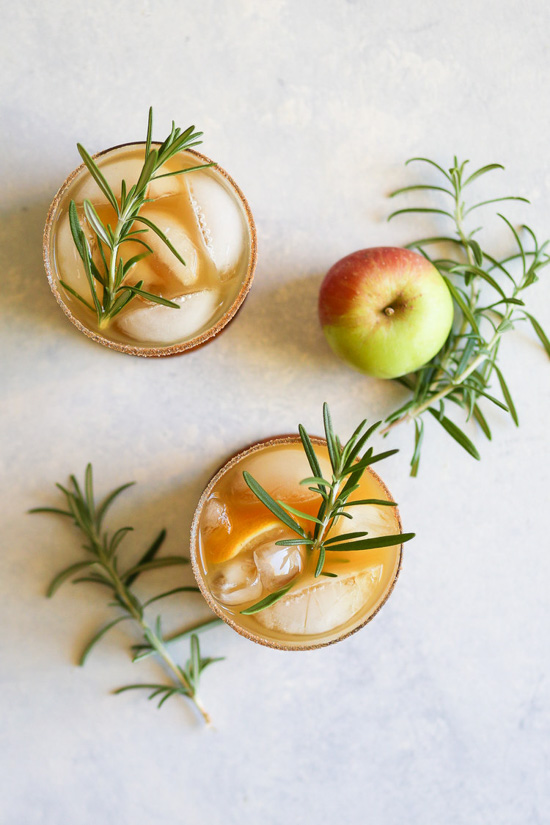 Aerial view of two cocktails in rocks glasses with rosemary sprigs placed around the glasses and an apple. 