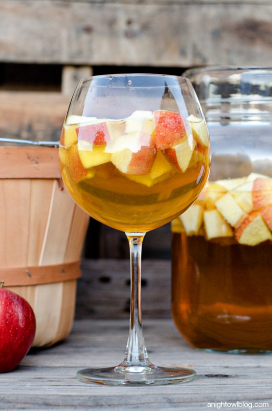 Large glass filled with apple sangria and a large jug of it in the background next to an apple picking basket. 