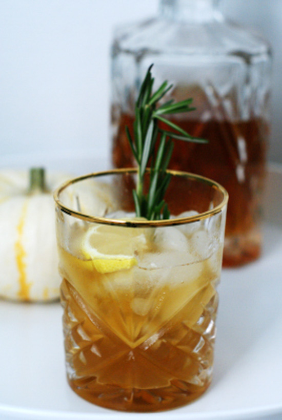 An etched tumbler with a gold-rim filled with a cocktail and a lemon and rosemary garnish with a decanter in the background. 