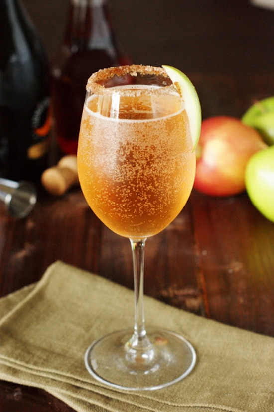 White wine glass with a sugared-rim, filled with sparkling peach colored liquid placed on a linen napkin with apples in the background. 