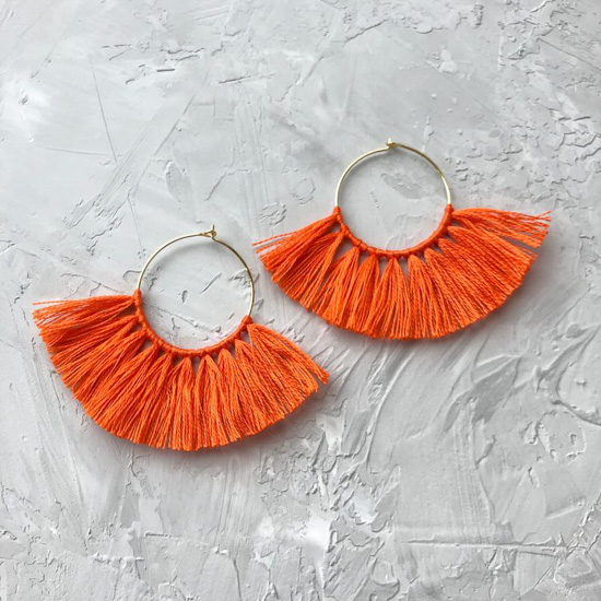 Halloween orange colored tassel earrings on a gold hoop base, laid flat on a textured piece of concrete. 