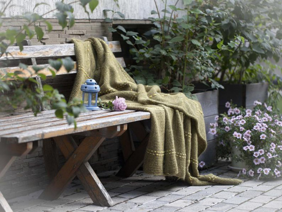 Hand-knit blanket in a muted shade of olive green laid over a rustic wooden bench and surrounded by foliage.