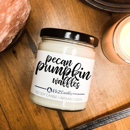 White candle in a glass container with a black lid in the scent 'pecan pumpkin waffles' placed on a wooden table with a burning candle next to it. 