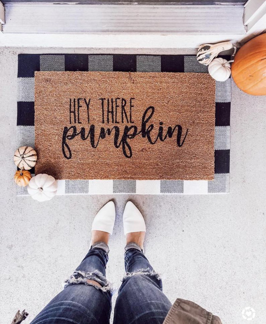 Halloween decor, jute rug with 'hey there pumpkin' written on it and laid over a black and white buffalo check surrounded by pumpkins and female legs wearing jeans and white flats.