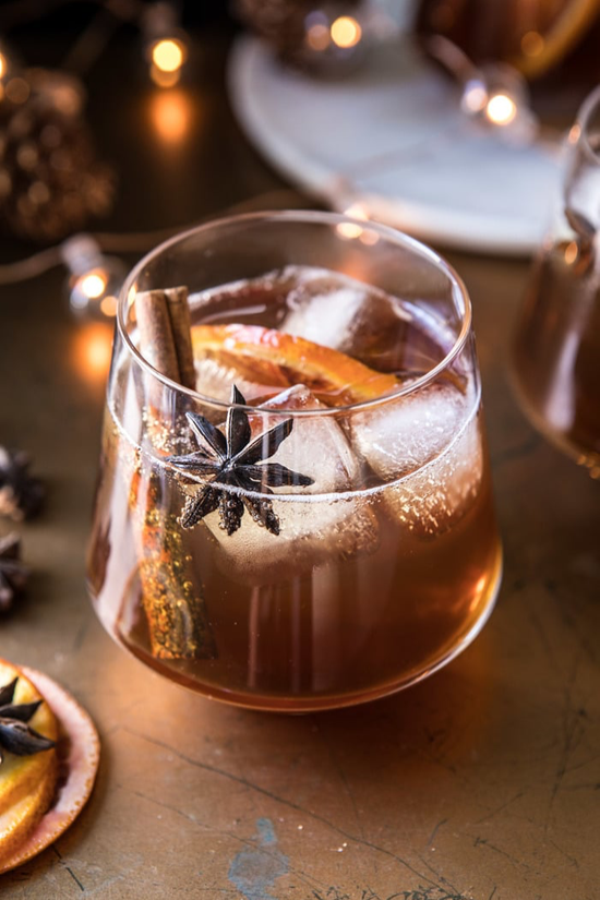 Fancy stemless glass filled with a caramel colored cocktail and garnished with an orange slice cinnamon stick.