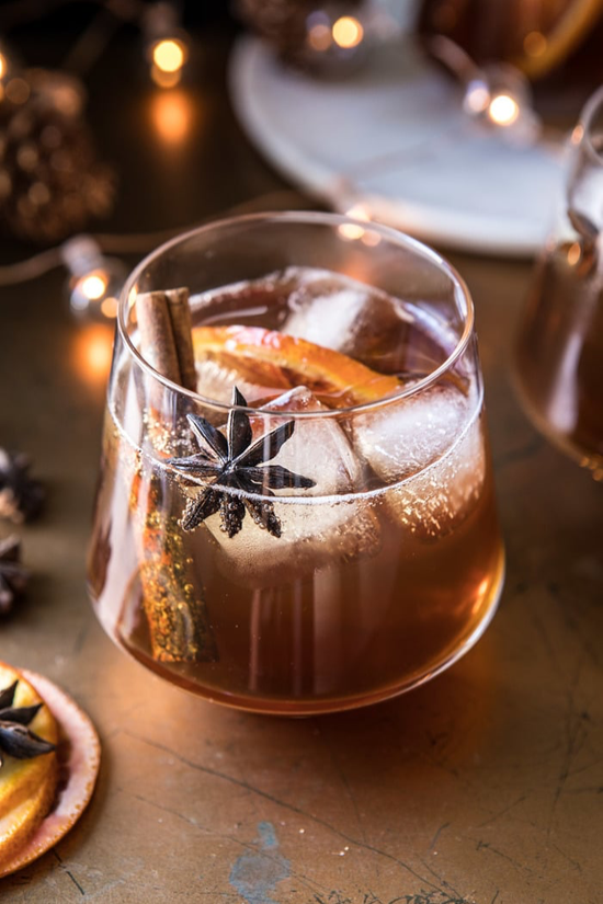 Fancy stemless glass filled with a caramel colored cocktail and garnished with an orange slice cinnamon stick. 