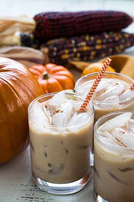 Three frothy cocktails in tumblers with orange and white straws, pumpkins and multi-colored corn in the background.