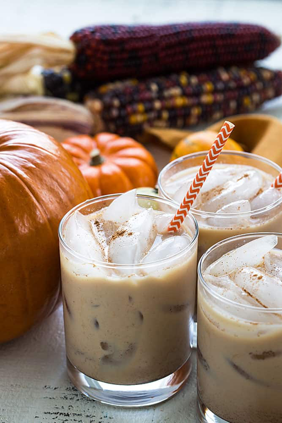 Three frothy cocktails in tumblers with orange and white straws, pumpkins and multi-colored corn in the background. 