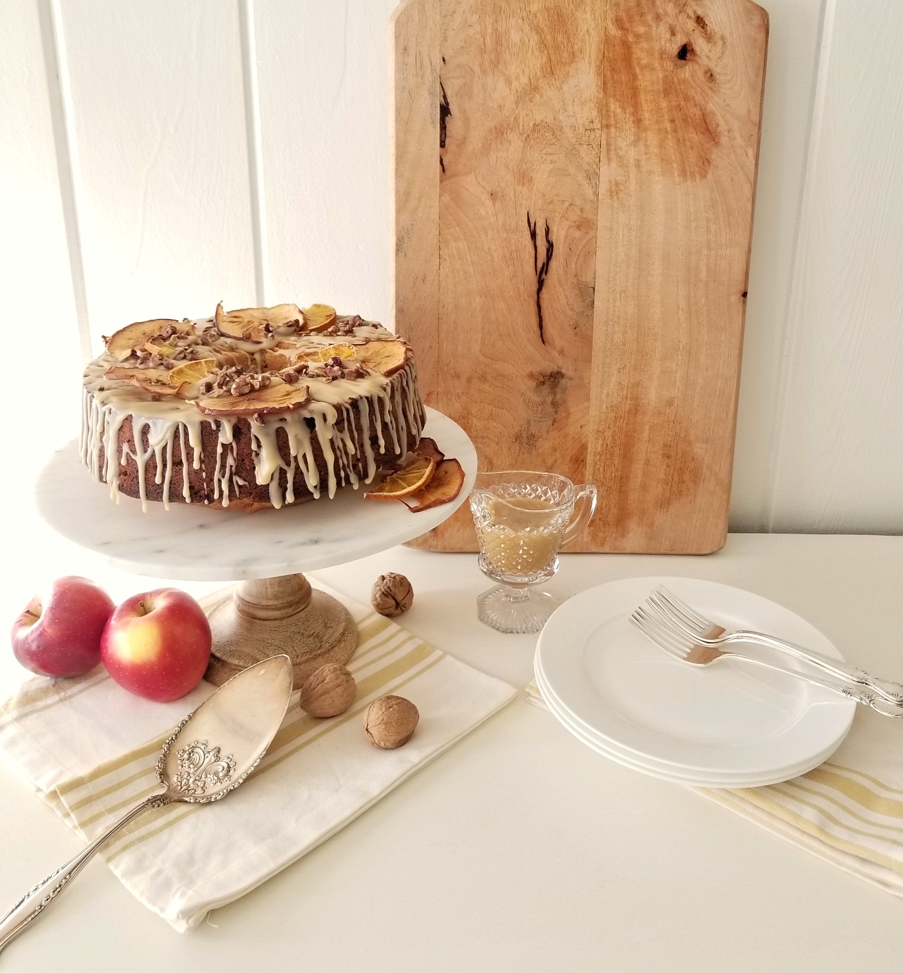 Apple spice cake on a marble cake stand adorned with a dripping glaze frosting and dries pieces of orange and apple, shiplap walls and a wooden cutting board in the background. 