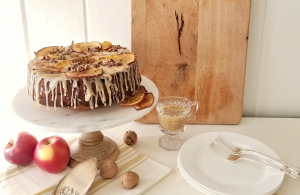Apple spice cake on a marble cake stand adorned with a dripping glaze frosting and dries pieces of orange and apple, shiplap walls and a wooden cutting board in the background.