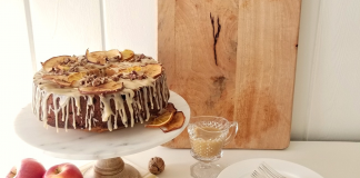 Apple spice cake on a marble cake stand adorned with a dripping glaze frosting and dries pieces of orange and apple, shiplap walls and a wooden cutting board in the background.