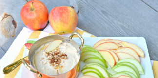 Horizontal image of a pan filled with caramel apple cream cheese dip on a white tray and surrounded by green and red apple slices.