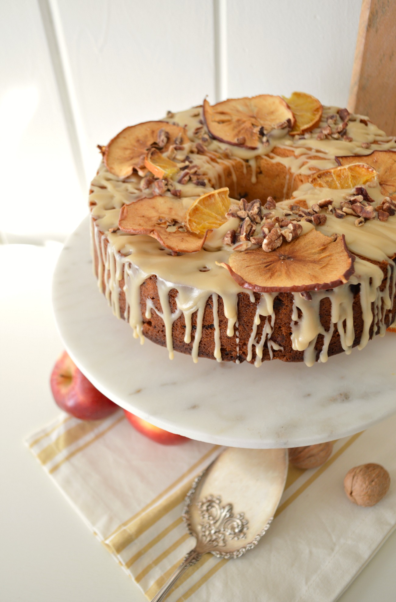 Apple spice cake on a marble cake stand adorned with a dripping glaze frosting and dries pieces of orange and apple. 