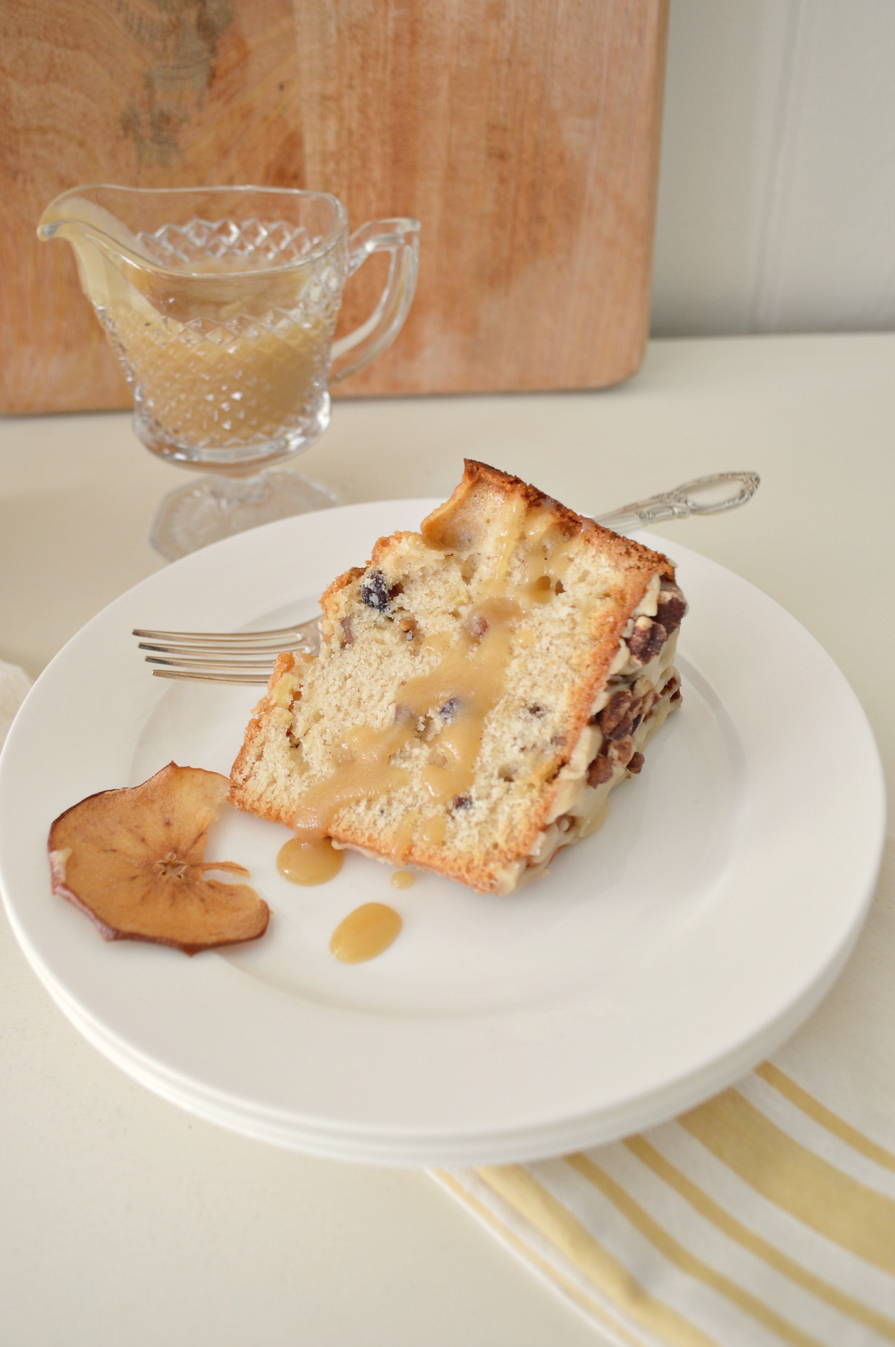 A single slice of apple spice cake topped with extra drizzle on a white glass plate with an ornate silver fork and a beveled glass gravy bowl filled with frosting. 