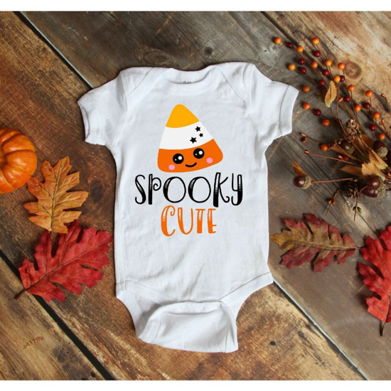 White baby onesie with a cartoon candy corn and "spooky cute" written on the front and placed on a wooden plank floor.