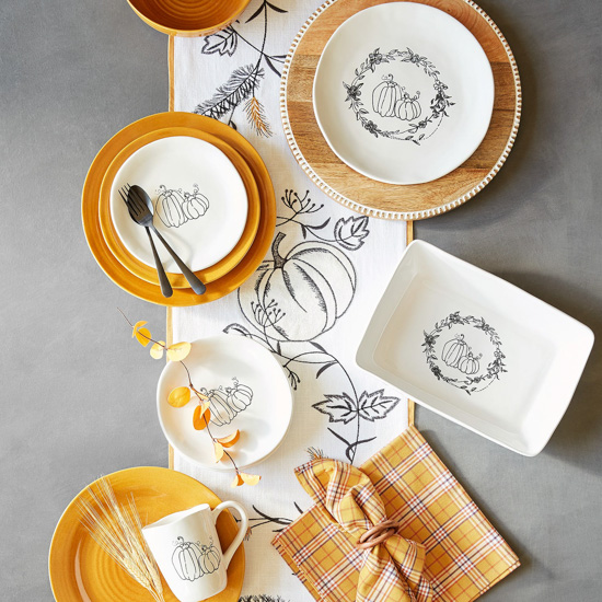 Aerial view of a display of Pier 1's Buffalo Harvest collection including mugs, plates and serve ware with a simplistic pumpkin line design on white dishes. 