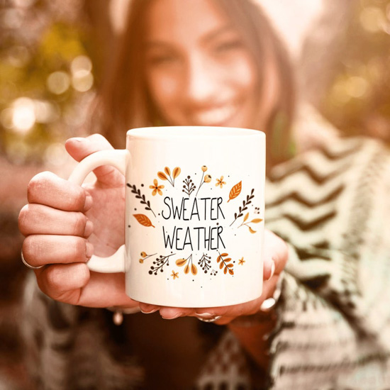 Girl in nature holding out a white mug that says "sweater weather" surrounded by fall colored leaves.