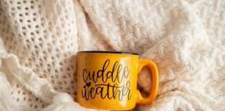 Cream colored blankets on the background and a ceramic, mustard colored, camp style mug that says "cuddle weather".