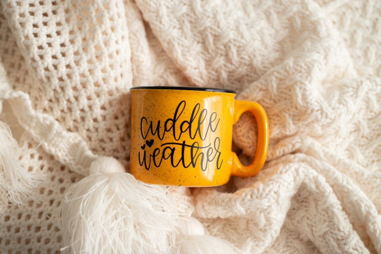 Cream colored blankets on the background and a ceramic, mustard colored, camp style mug that says "cuddle weather".