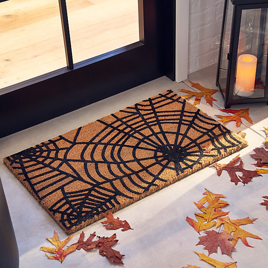 Jute welcome mat with black spiderwebs placed in front of a door and next to a lantern and fall leaves. 