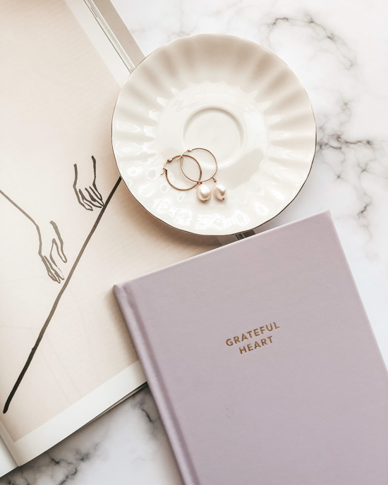 Pink hardcover journal that says "grateful heart" on the cover next to an open book and a porcelain dish with pearl earrings in it. 