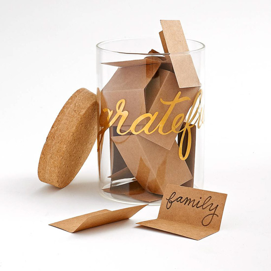 Glass jar with "grateful" written around the outside in gold calligraphy, filled with Kraft paper notes of thankfulness with a cork lid.