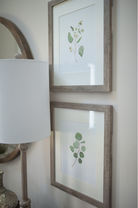 A lamp on a sideboard table with a white shade in front of two hung and framed pieces of art on the wall. 