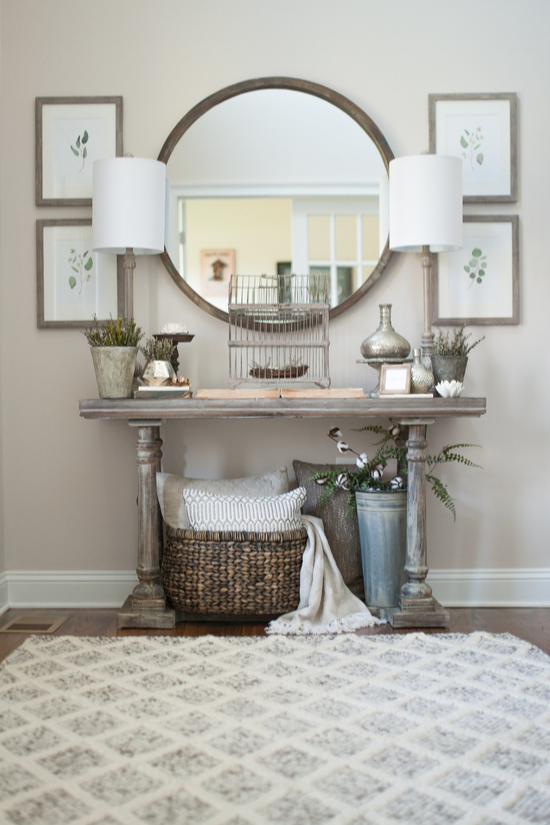 An inviting entryway, a cozy rug, end table with matching lamps, artwork to look at and large round mirror. 