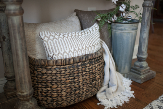 Large woven brown basket containing cozy white and oatmeal colored pillows and a throw blanket. 