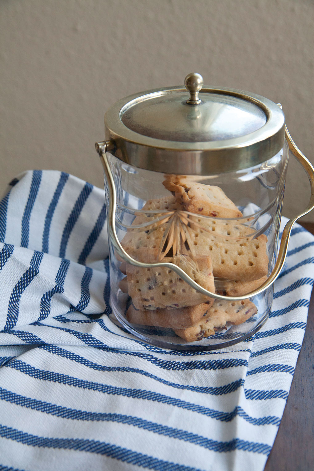 Glass biscuit jar filled with tea biscuits set out on a blue and white striped tea towel. 