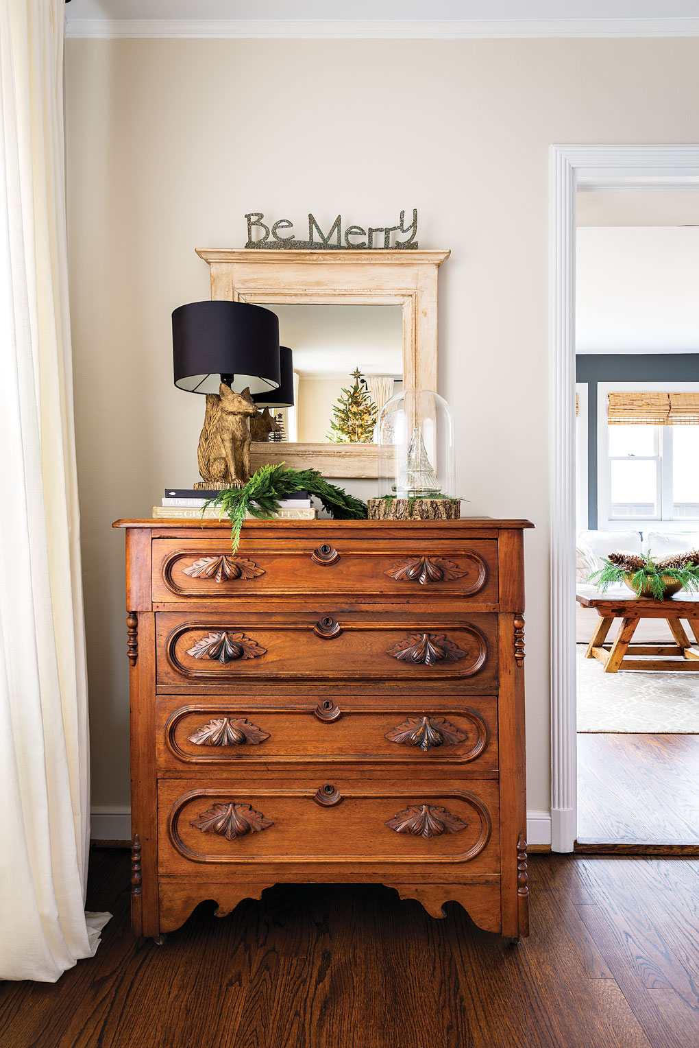 An antique and ornate wooden dresser with a mirror and a fox lamp on top accented with fresh greenery.