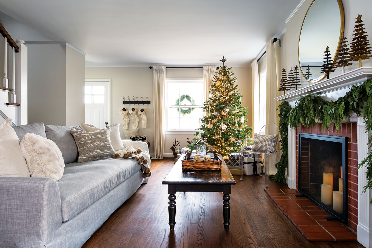 Wide shot into a simply lovely family room with light gray couch placed across from a candle-filled fireplace with a lighted Tree in the background and white stockings hung by the front door.