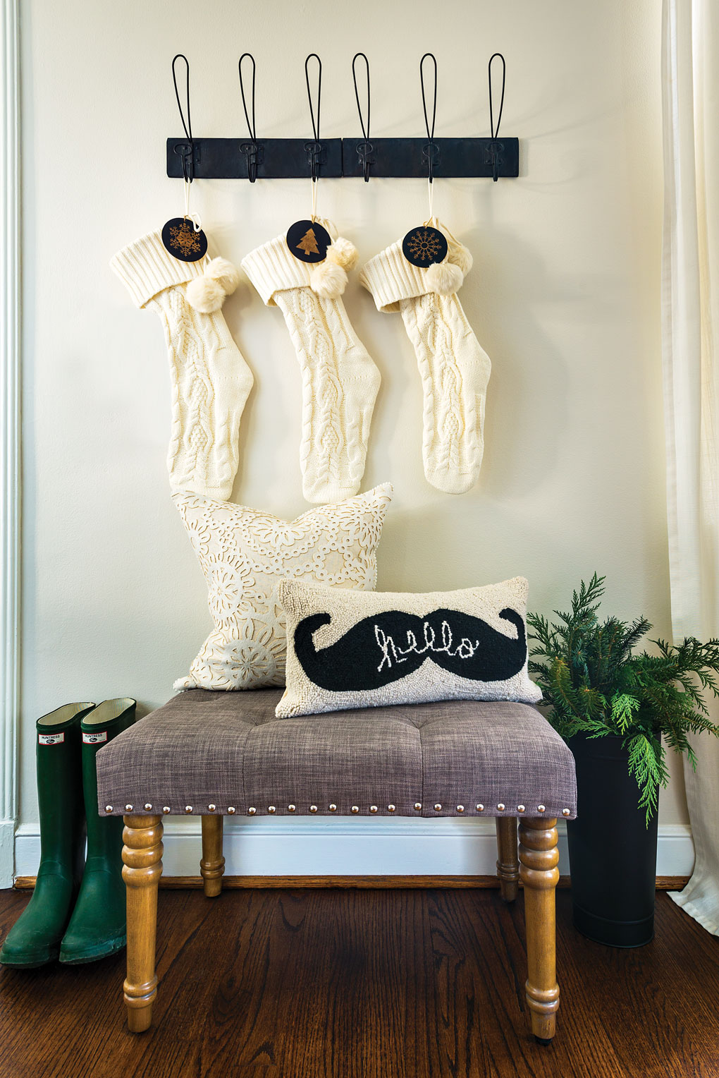 An entryway with a small grey bench next to green Hunter boot and fresh greenery with knitted cream colored stockings hanging overhead.