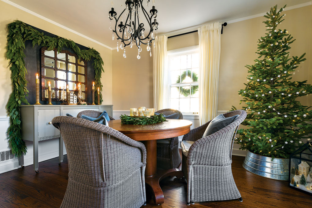 Wicker chairs in gray surround the dining table while a black and crystal chandelier centered above it. Real greenery in the form of a garland and a Christmas tree add color to the room. 