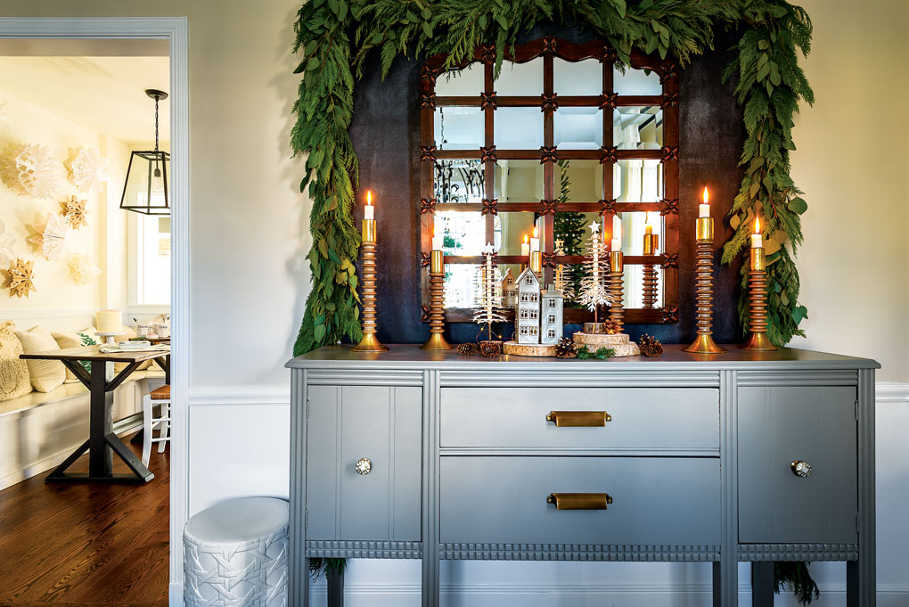 Light blue-gray buffet with a framed window mirror sitting upon it with lit tapered candles and greenery draped around the window. 