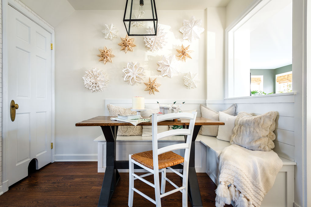 A white kitchen banquette with a wooden dining table and handmade paper snowflakes hanging on the wall. 