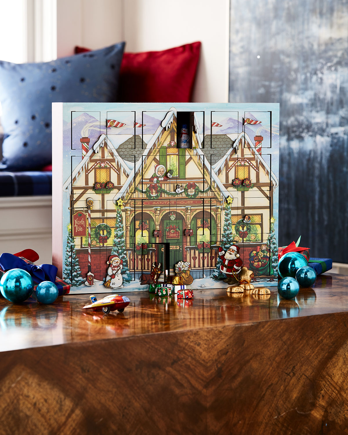 Wooden advent calendar filled with tiny ornaments, surrounded by glass ornaments and sitting on a wooden table.