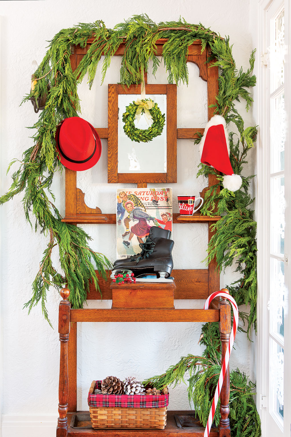 A wooden hall tree in the entryway of the home adorned with a cedar garland with pops of red and a pair of antique black ice skates. 