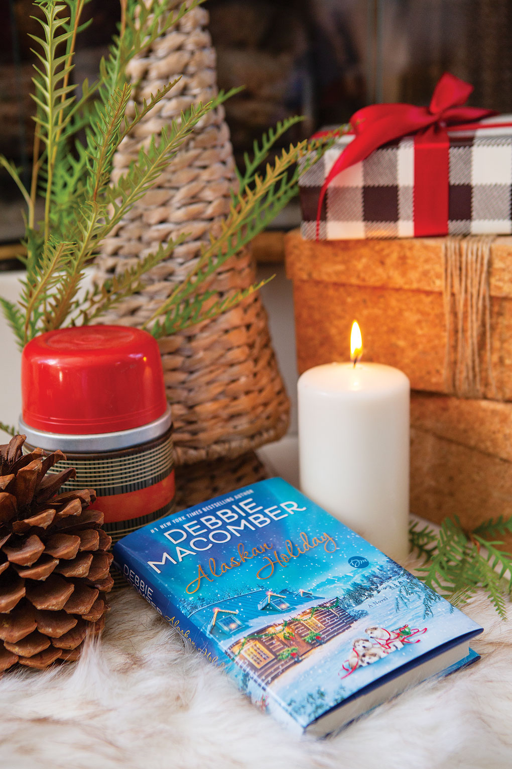 Vignette featuring a rustic Christmas themed book surrounded by pinecone, thermos, candle and stacked presents. 
