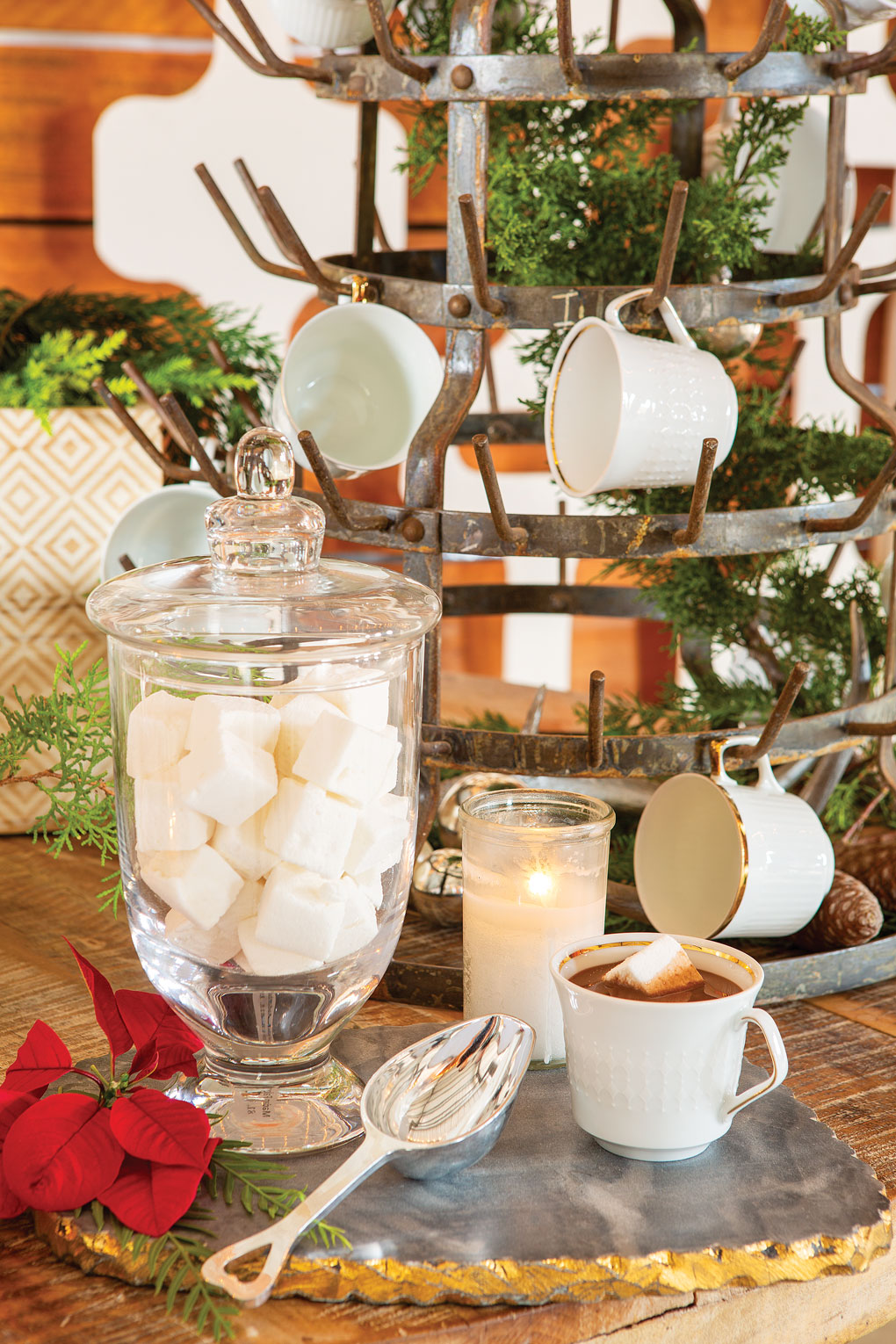 Glass apothecary jar filled with large marshmallows cubes next to a large metal mug holding tree and a cup of cocoa. 