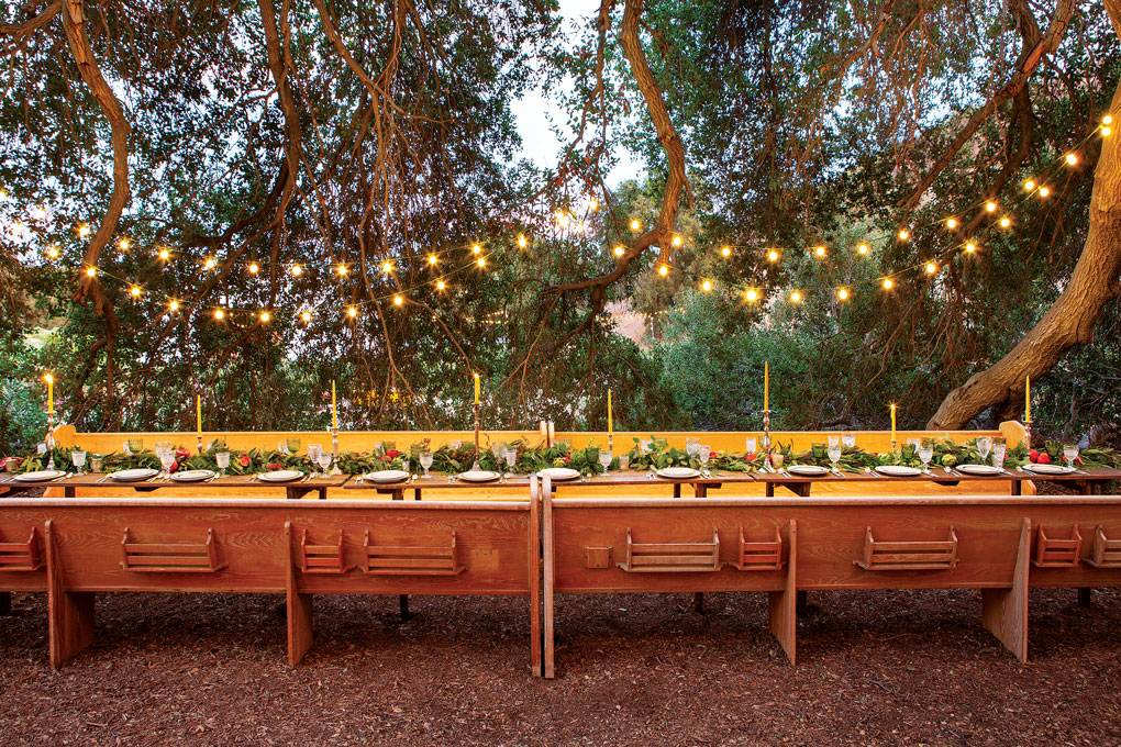 Festive farm outdoor dining setup in the form of a long family style table made up of farm tables and church pews with string lights hanging from the trees above. 