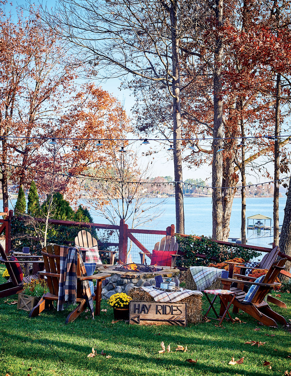 Adirondack chairs encircle a fire pit next to the lake, surrounded by trees and string lighting. 