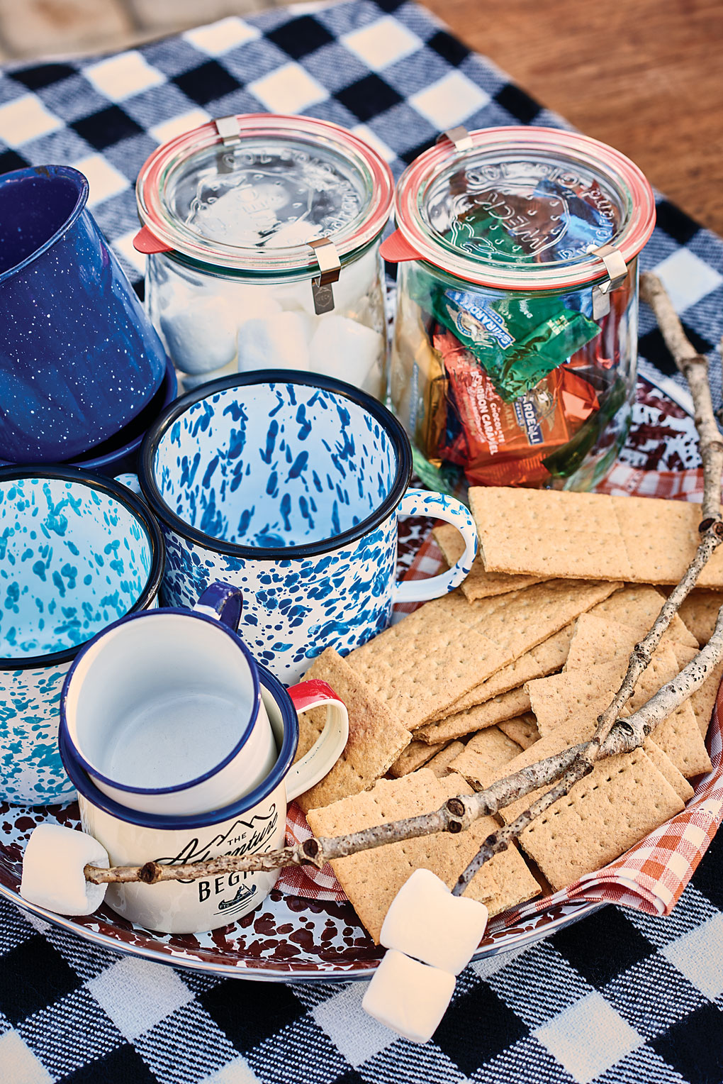 Ceramic, speckled camp mugs laid out with marshmallows, graham crackers and chocolates for a hot cocoa and s'mores station near the fire pit. 