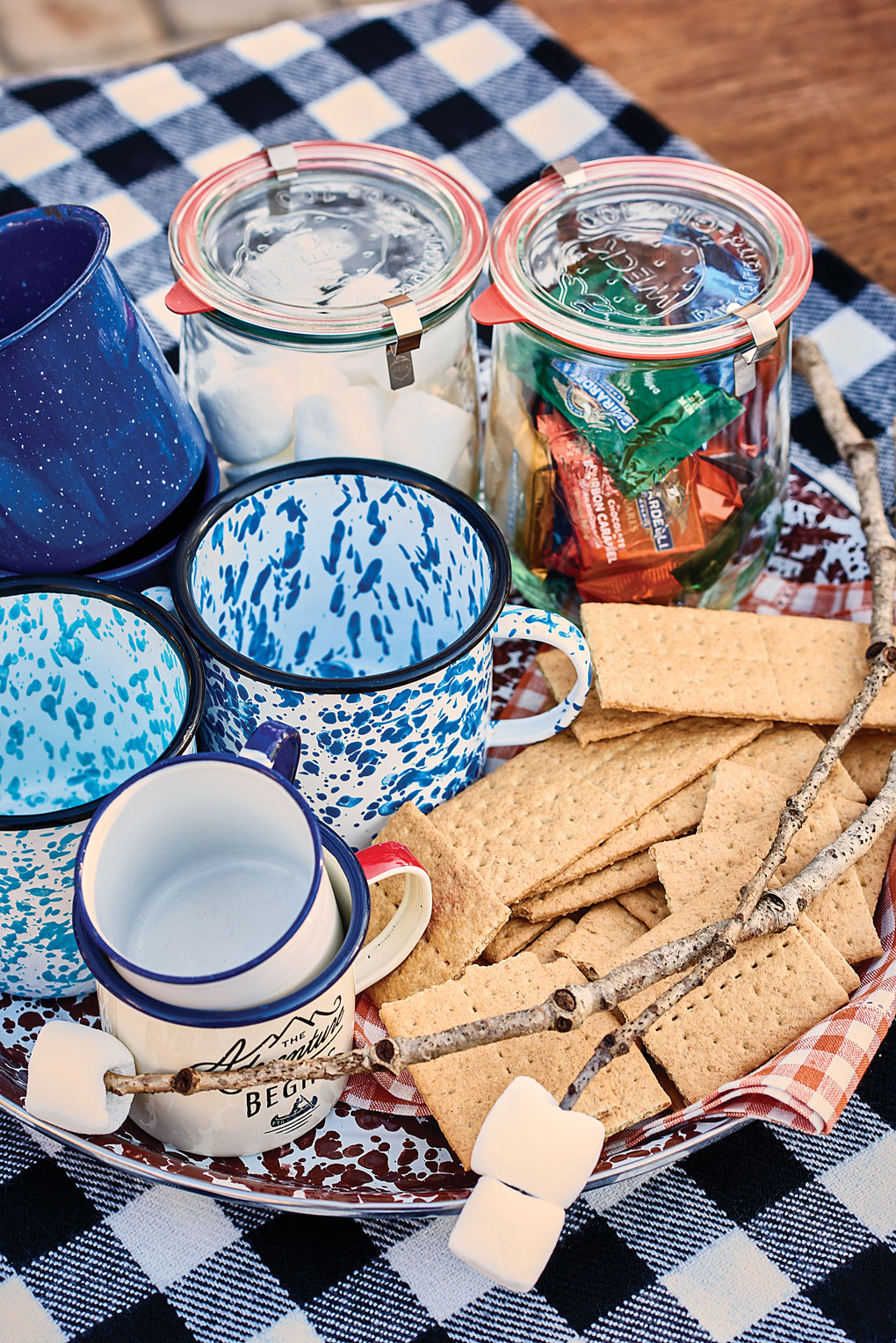 Ceramic, speckled camp mugs laid out with marshmallows, graham crackers and chocolates for a hot cocoa and s'mores station near the fire pit.