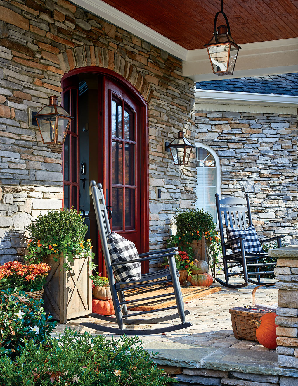 Stonework facade and deep red double front doors, flanked by black wooden rocking chairs and scattered pumpkins for a fall welcome. 